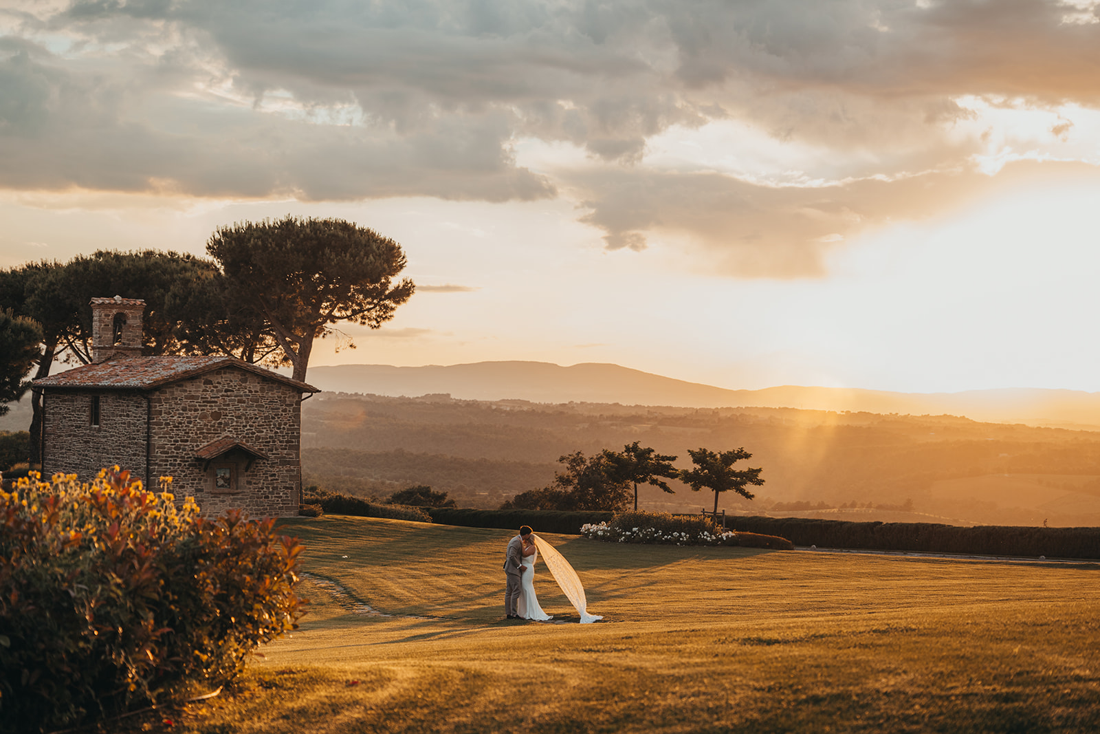 Landscape photo of a couple walking upon a hill at sunset, surrounded by rolling hills, with the bride and groom in the distance, the brides veil blowing in the evening wind