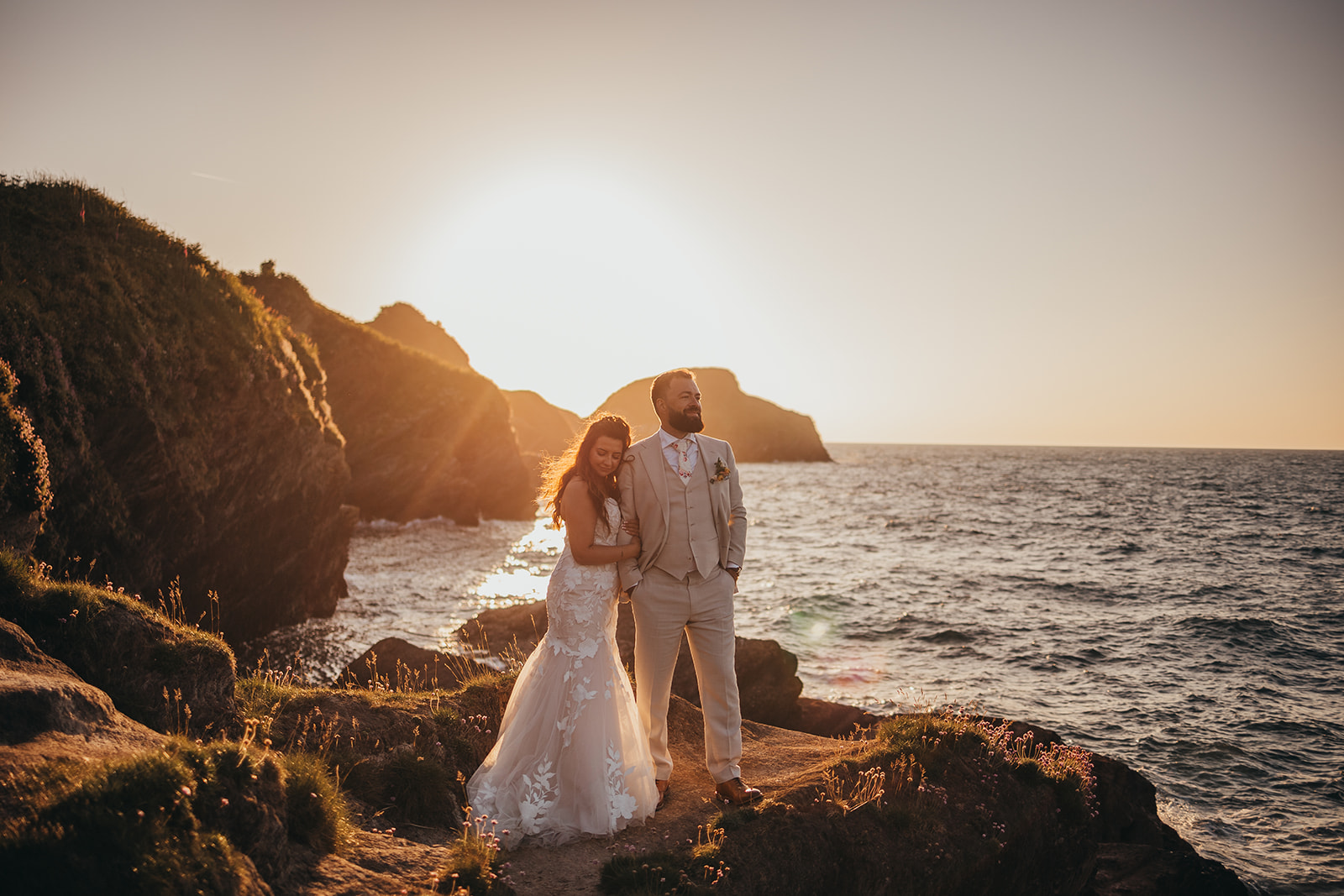 Bride and Groom, stood upon a rock on the cliffs over looking the sea, sea breeze in the air as the sun sets, casting a golden hue across the land.